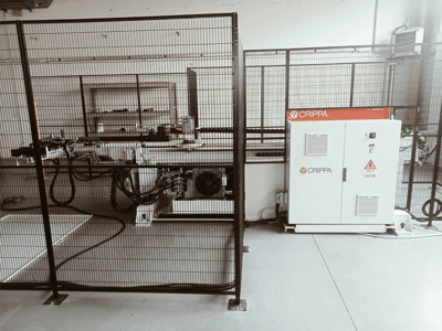 White CRIPPA machine behind a safety cage, with visible hoses and components, side view, industrial setting.