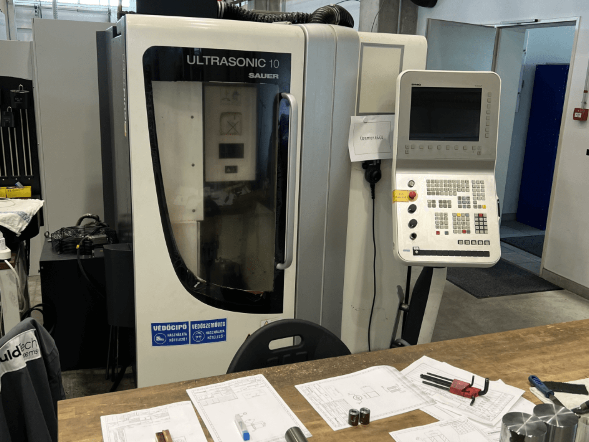Front view of DMG Ultrasonic 10 vertical machining centre with control panel, showing design documents and tools on a table.