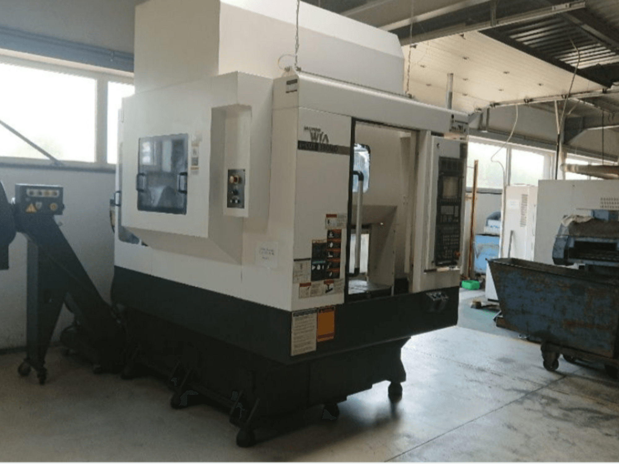 Haas CNC machine, side view, with control panel, warning labels, and a loading chute, set in a workshop environment.