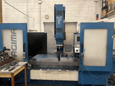A front view of a JAGIMA CNC machine with a large metal workspace, visible control panel, and tools on a nearby table.