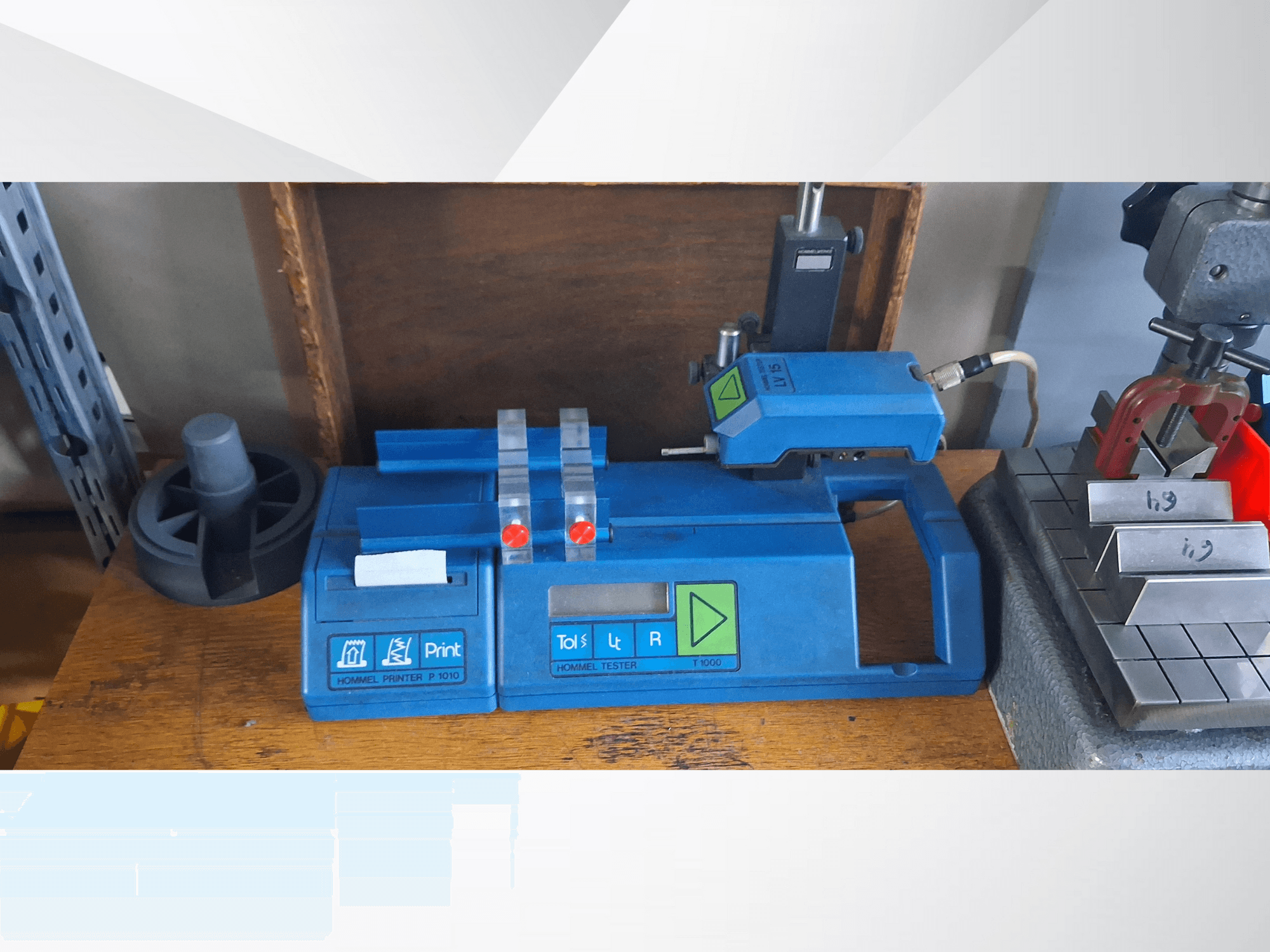 Blue Hommel T 1000 printer and T 1 010 tester with red buttons, viewed from the front on a workbench with metal blocks.