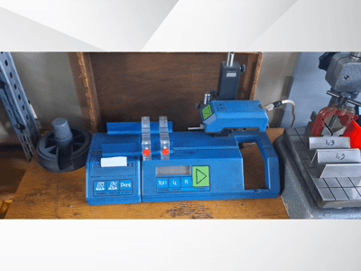 Blue Hommel T 1000 printer and T 1 010 tester with red buttons, viewed from the front on a workbench with metal blocks.