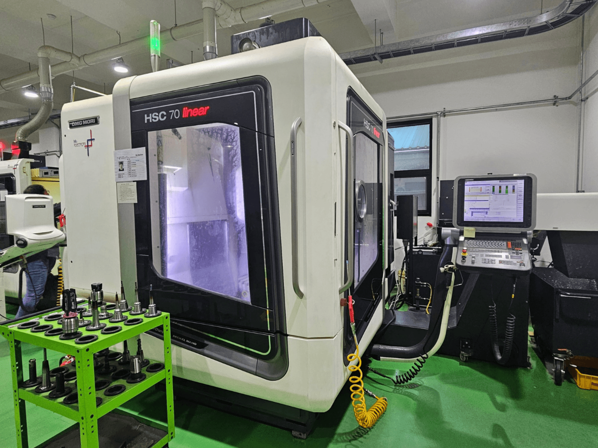 DMG MORI HSC 70 linear universal machining center, front view, with tool holders on a green table and a control panel visible.