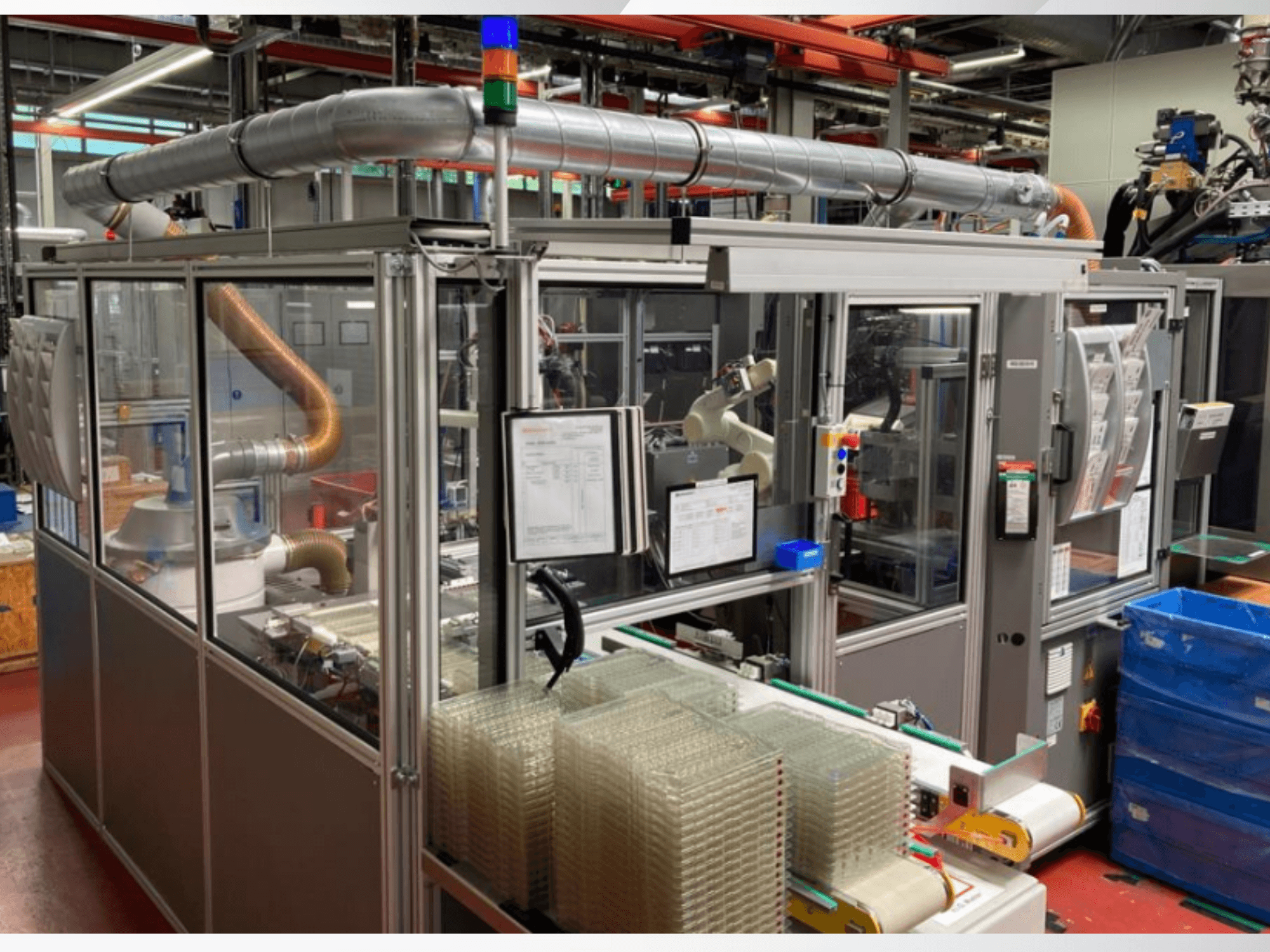 Front view of an automated machine with metal pipes, control panel, and stacked clear plastic containers on a factory floor.