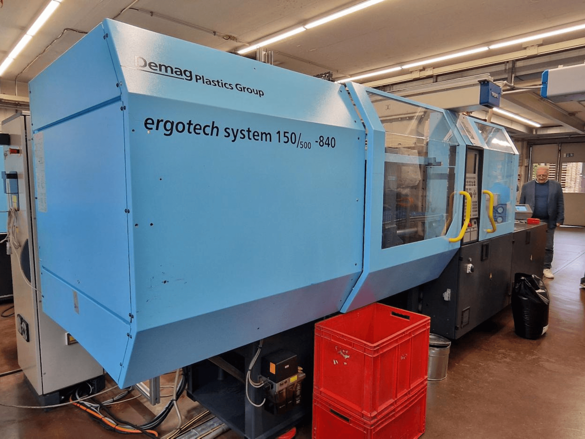 Demag Ergotech System 150/500-840 injection molding machine in blue, side view, with crates and operator in background.