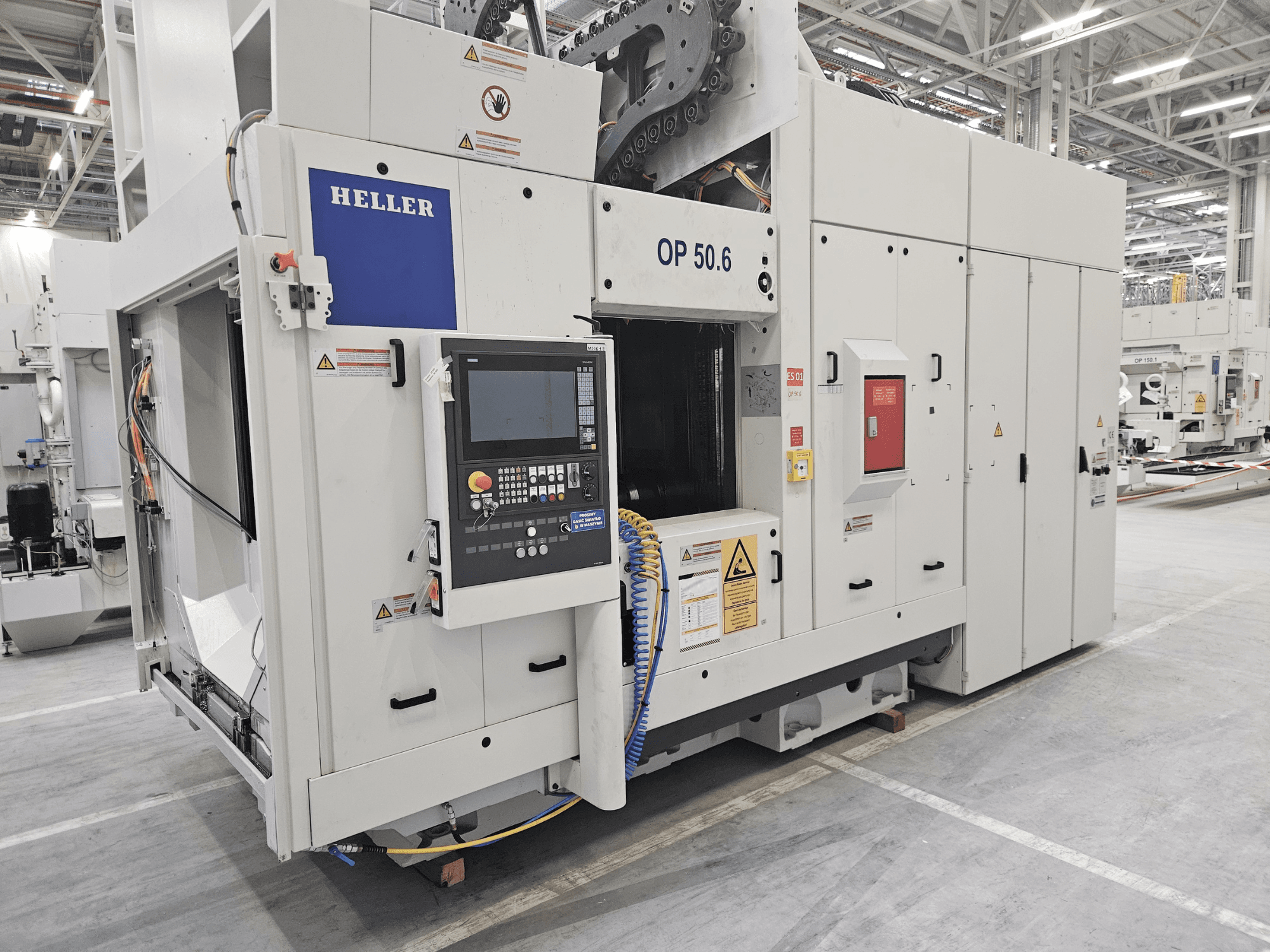 Heller OP 50.6 machining center, front view, with a control panel, warning labels, and hoses visible in a factory setting.