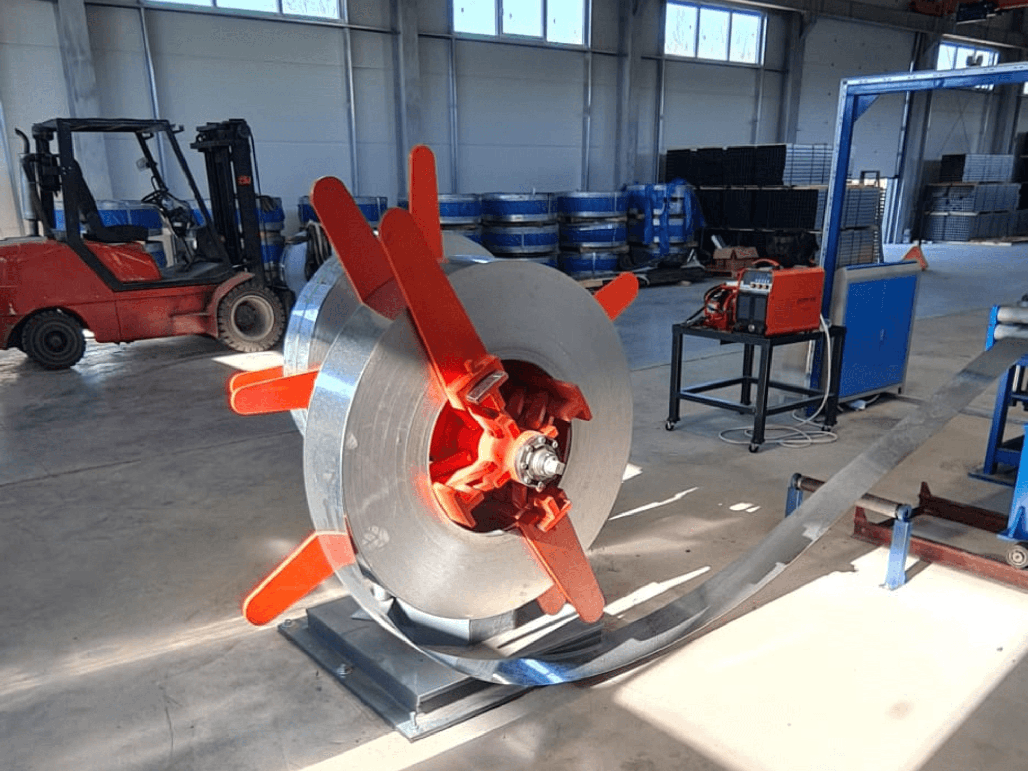 Metal coil with red handles on a stand, viewed from an angle. Industrial background with machinery and a forklift.
