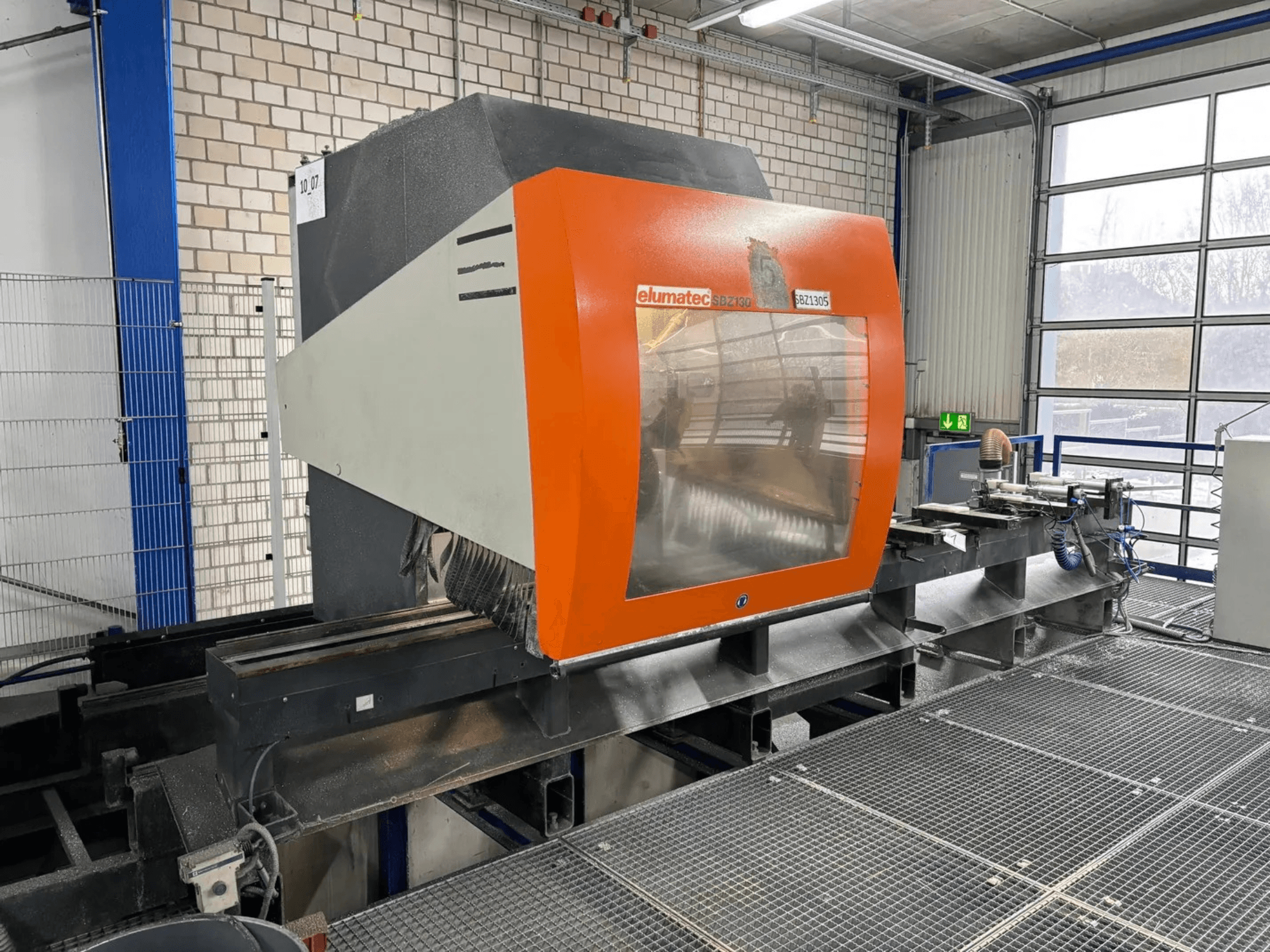 Elumatec SBZ 130 vertical machining center, front view, featuring an orange and gray housing with a transparent panel.