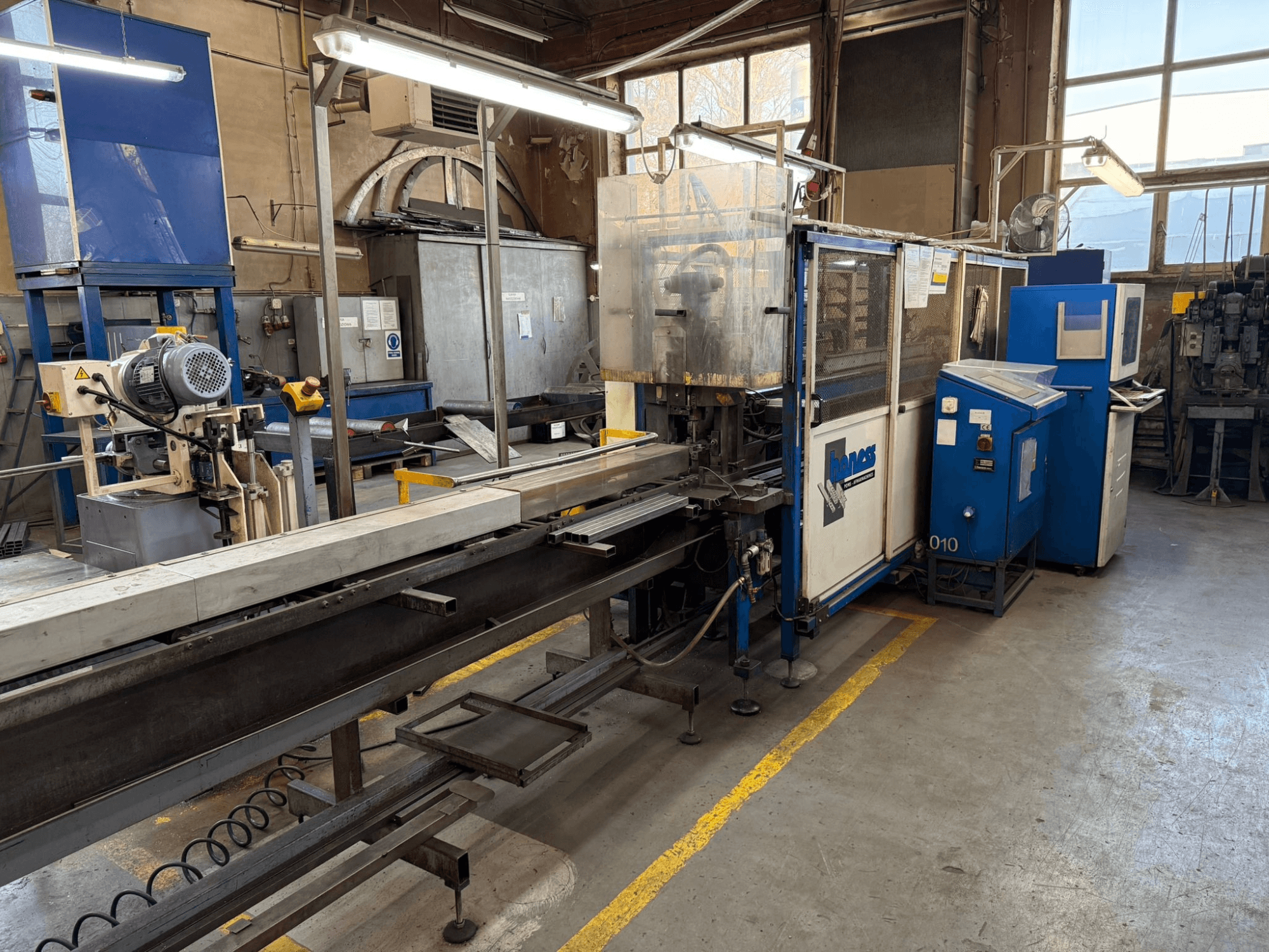 Industrial workshop with a blue Haweas machine, side view showing conveyor and equipment in a large, well-lit space.