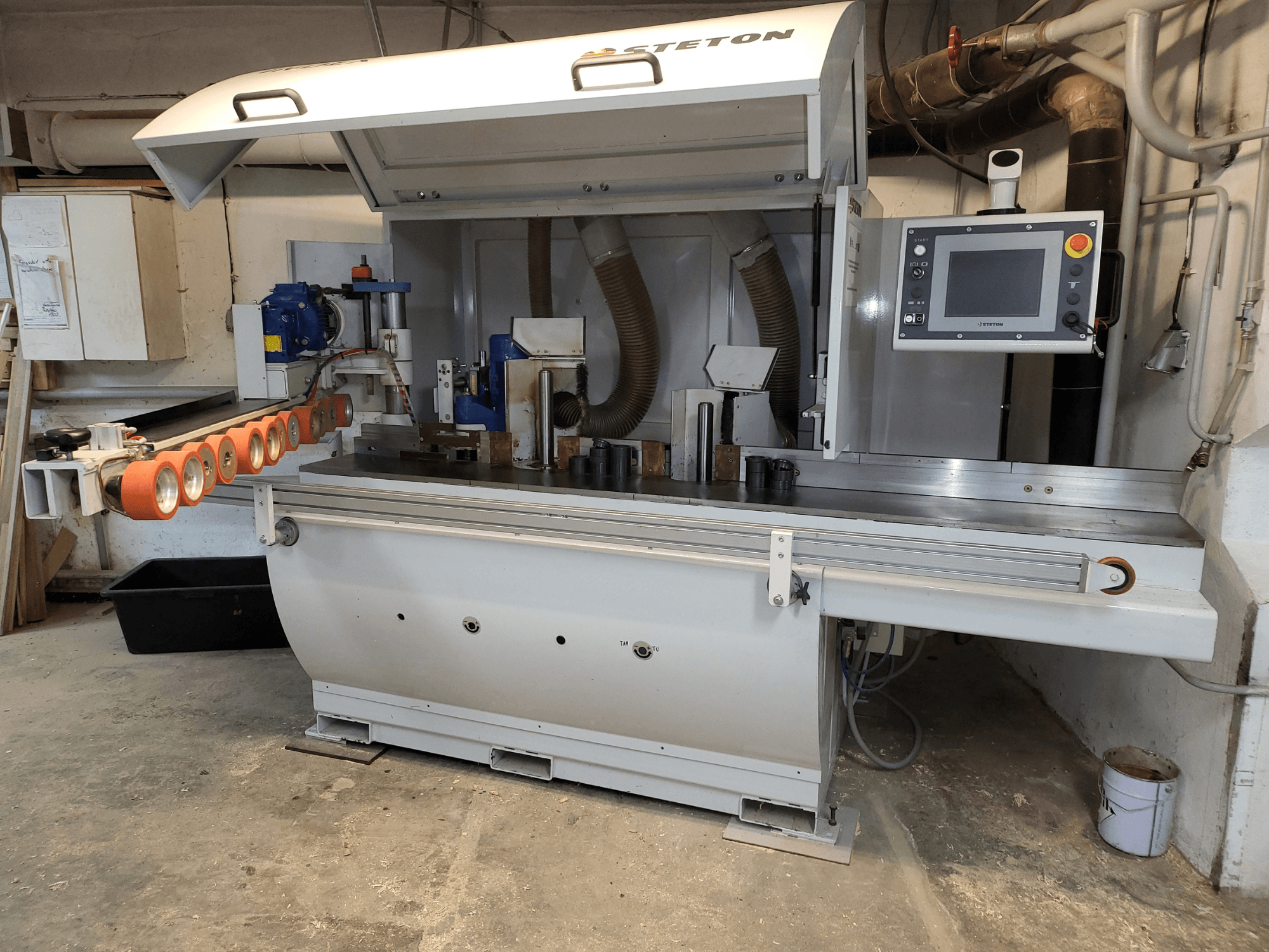 A front view of a Stetton woodworking machine with a control panel, feeder tubes, and various wood pieces on the table.