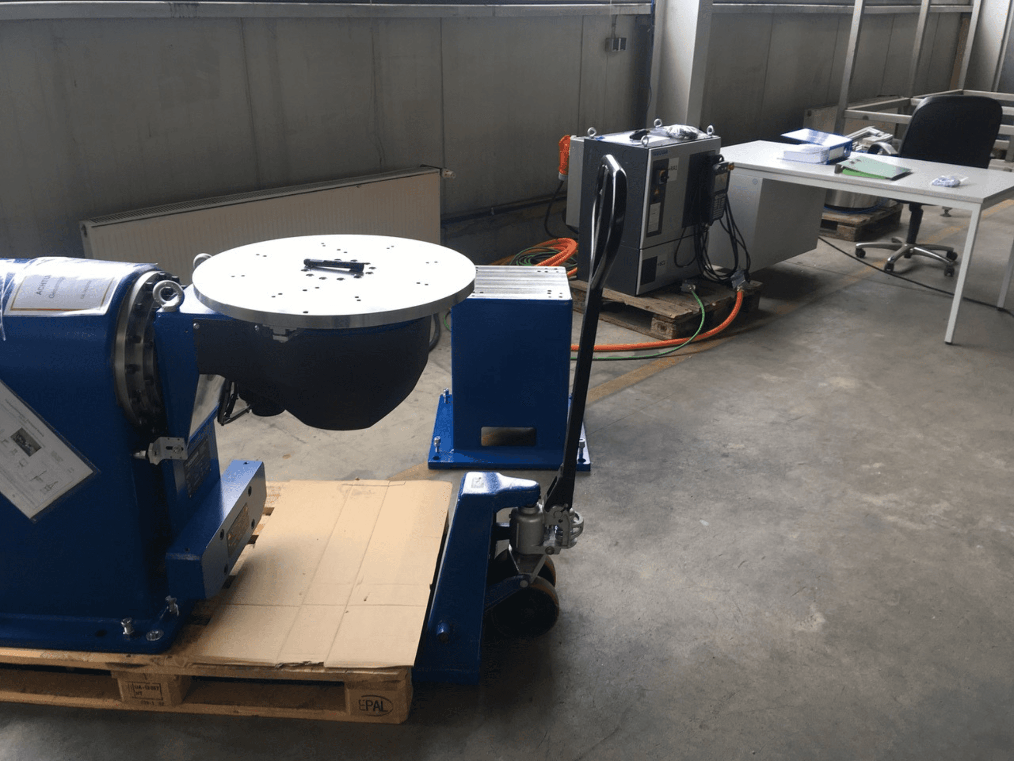 Blue industrial equipment with a circular base and control panel, viewed from the front, on a wooden pallet in an office setting.