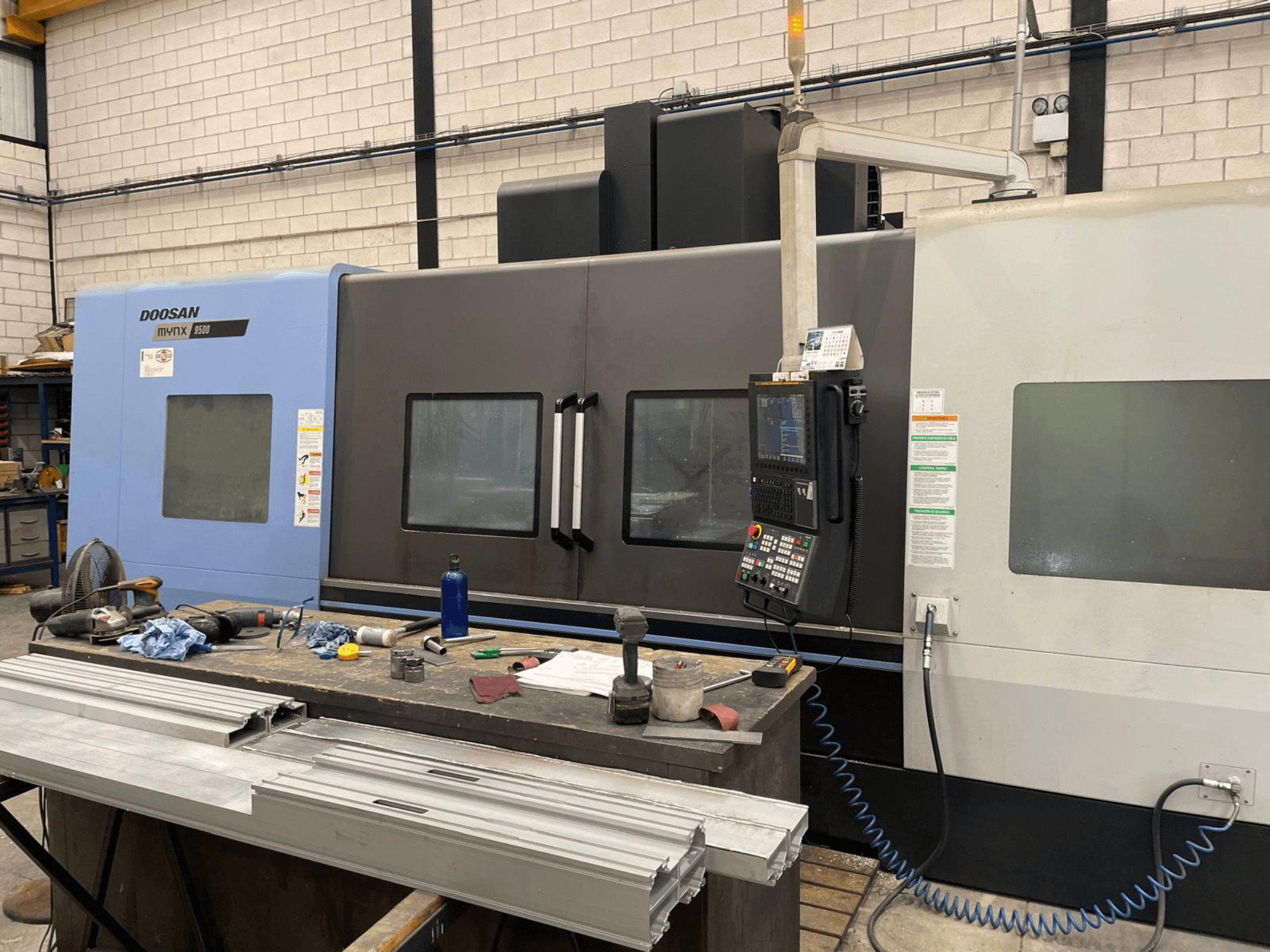 Doosan Mynx 6500 CNC machine in blue and gray with control panel, side view, workspace with tools and materials in foreground.