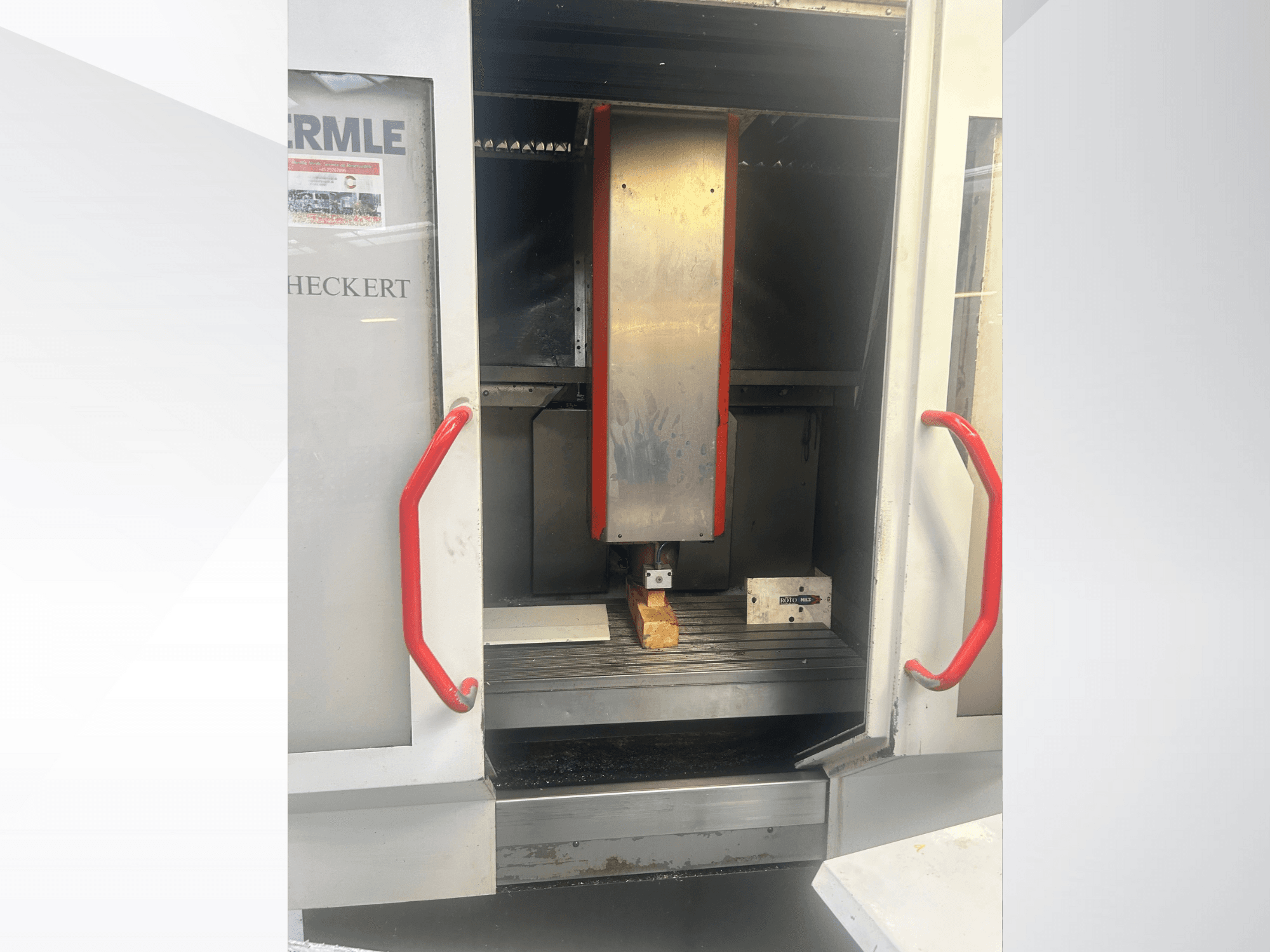 Hermle CNC machine front view, featuring a metallic interior, red handles, and a workpiece on the table.