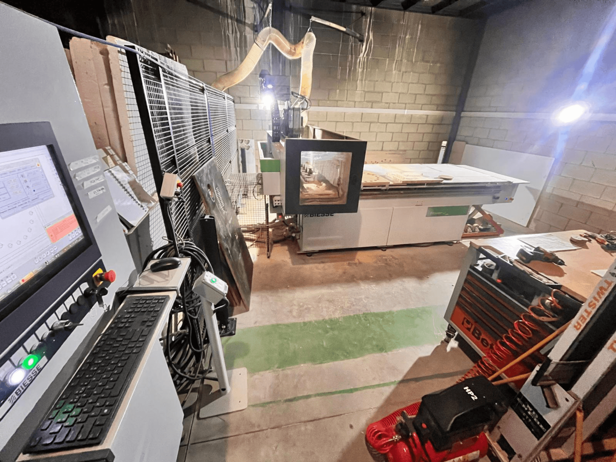 Biesse CNC machine in a workshop, side view showing control panel, dust extraction, tools, and various materials on the table.