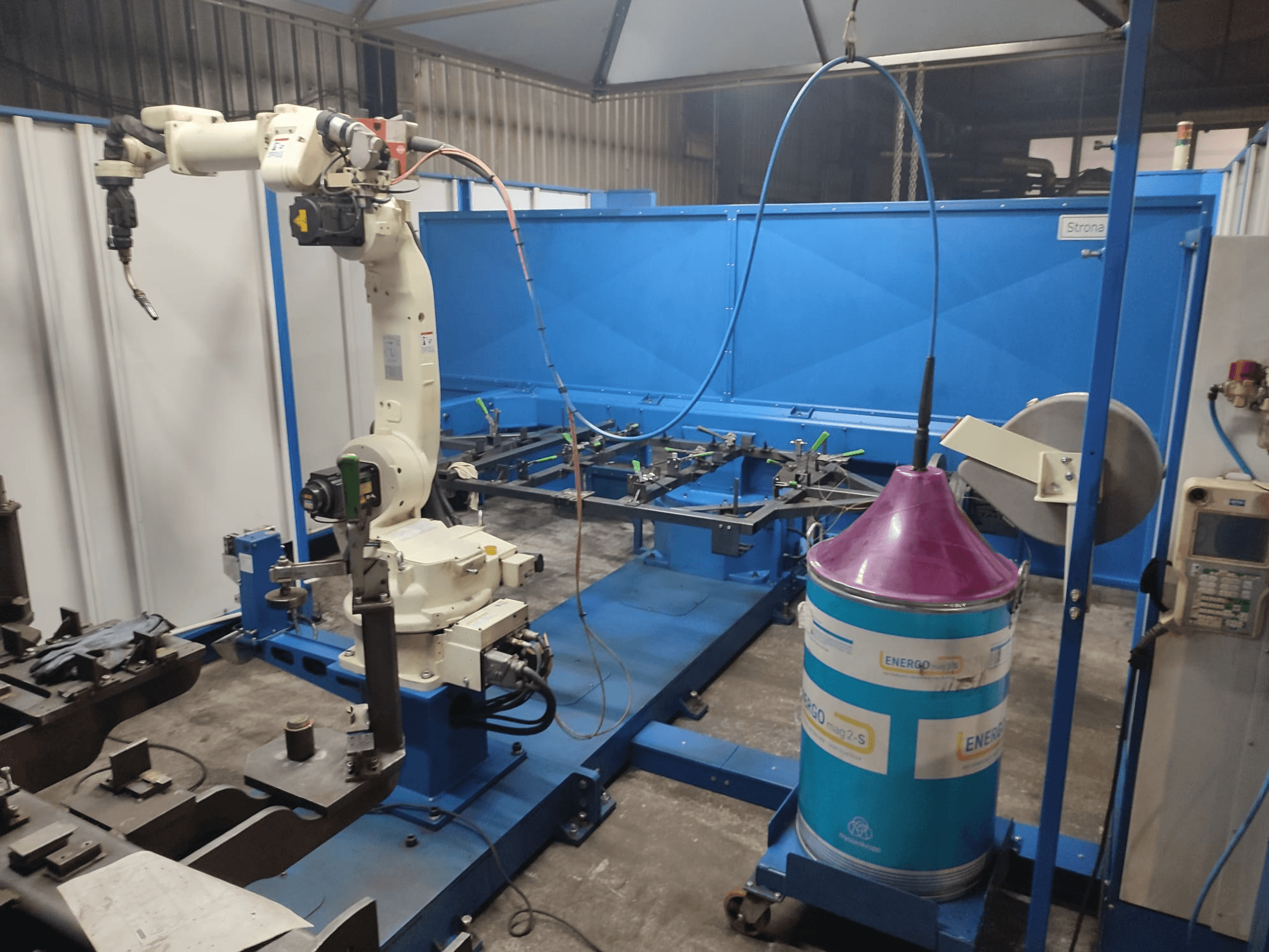 Industrial robotic arm with a white and blue base, setup in a workshop with a blue work surface and metallic components.