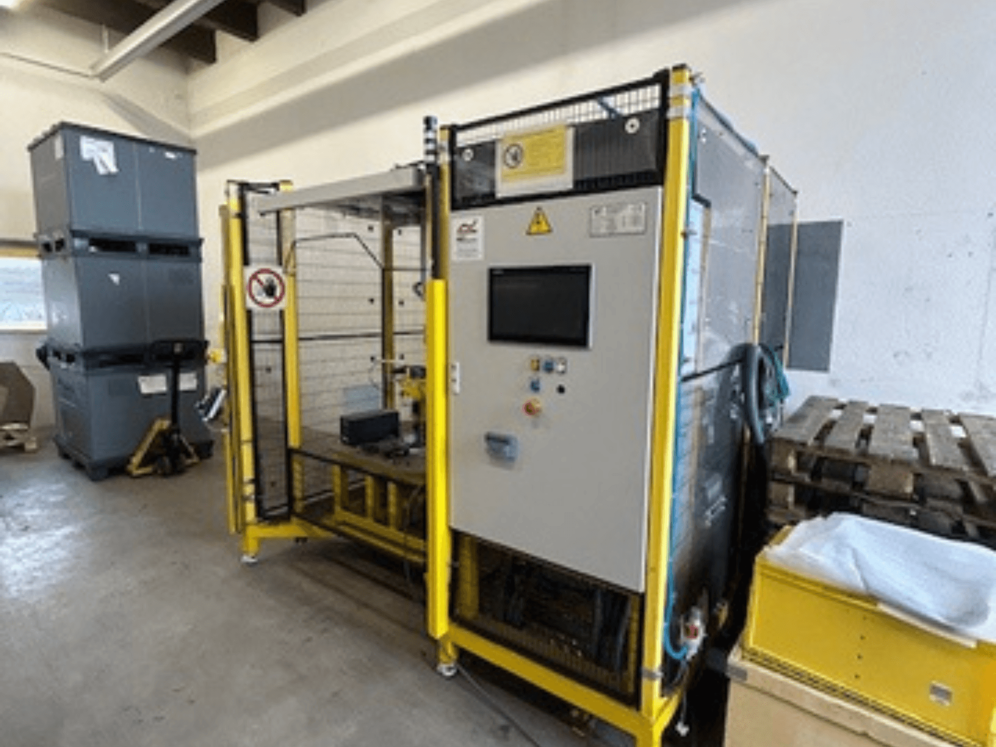 Industrial machine with a control panel and safety features, front view; yellow frame, grey bins, and pallets in background.