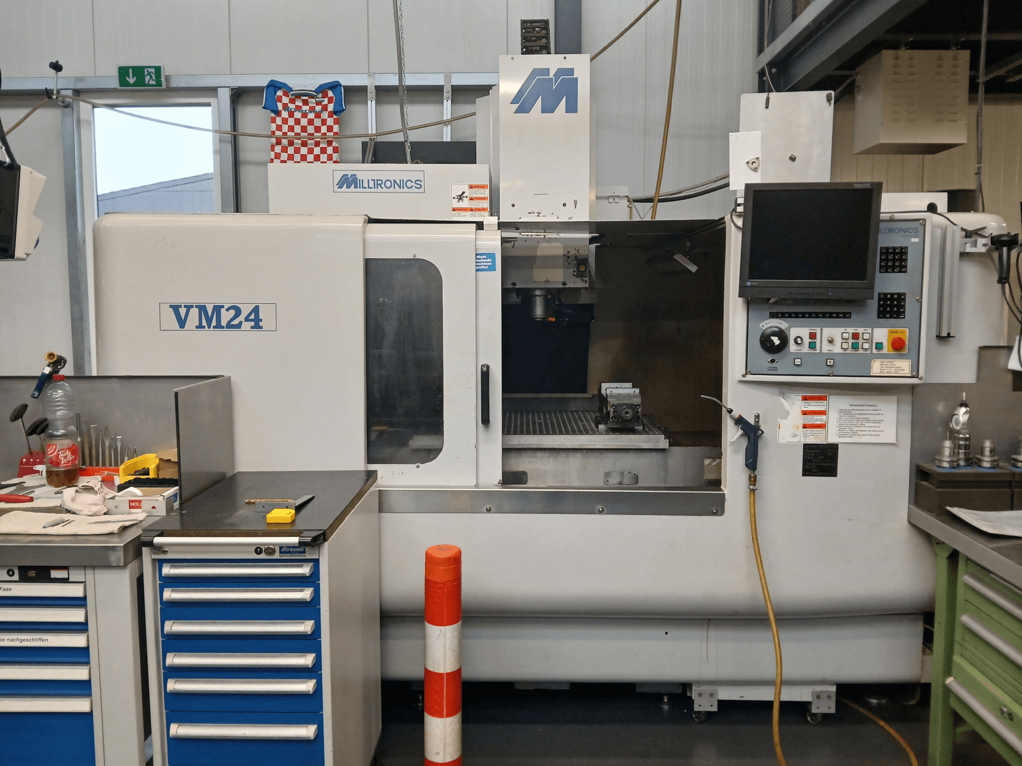 Milltronics VM24 CNC milling machine shown from the front, featuring a control panel, tool storage, and work area setup.