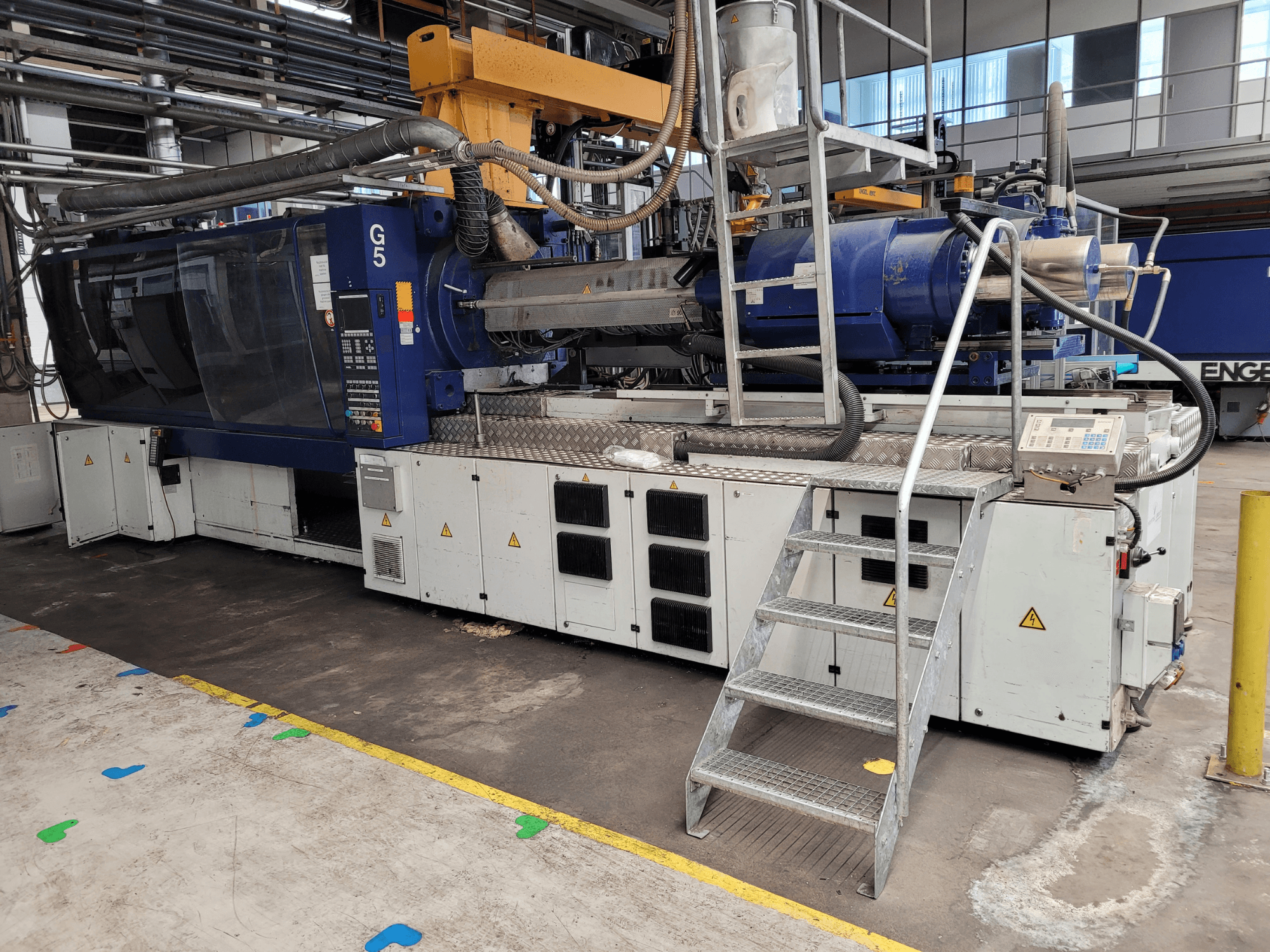 Injection molding machine G5, side view, with control panel, step ladder, and stainless steel work surface visible.