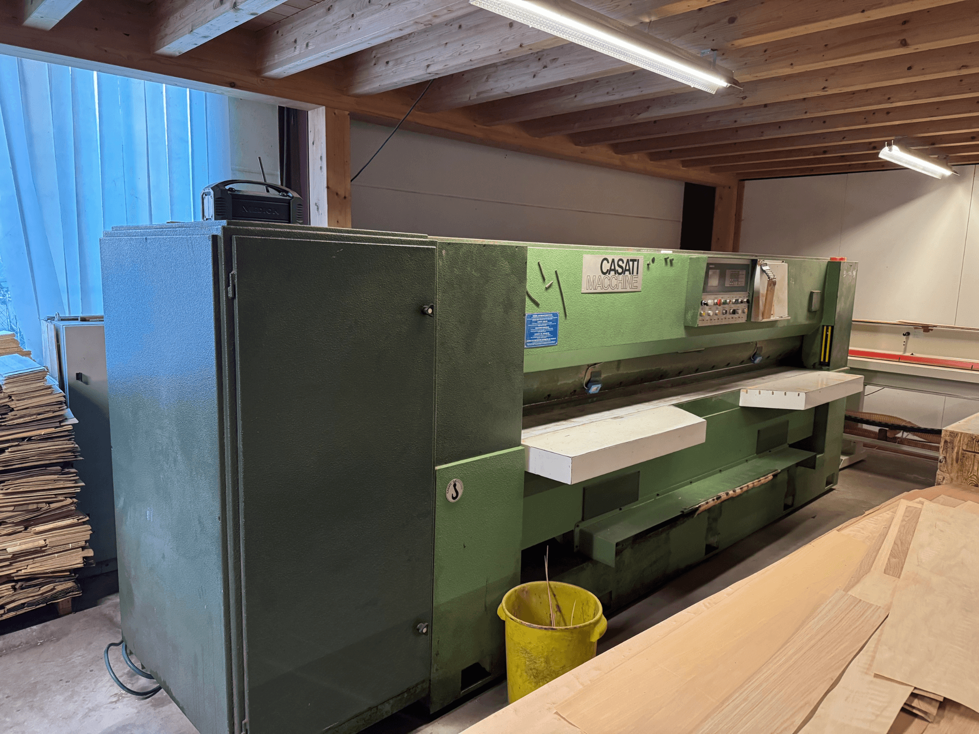 Green CASATI woodworking machine in a workshop, front view, with control panel and white work surfaces, surrounded by wood piles.