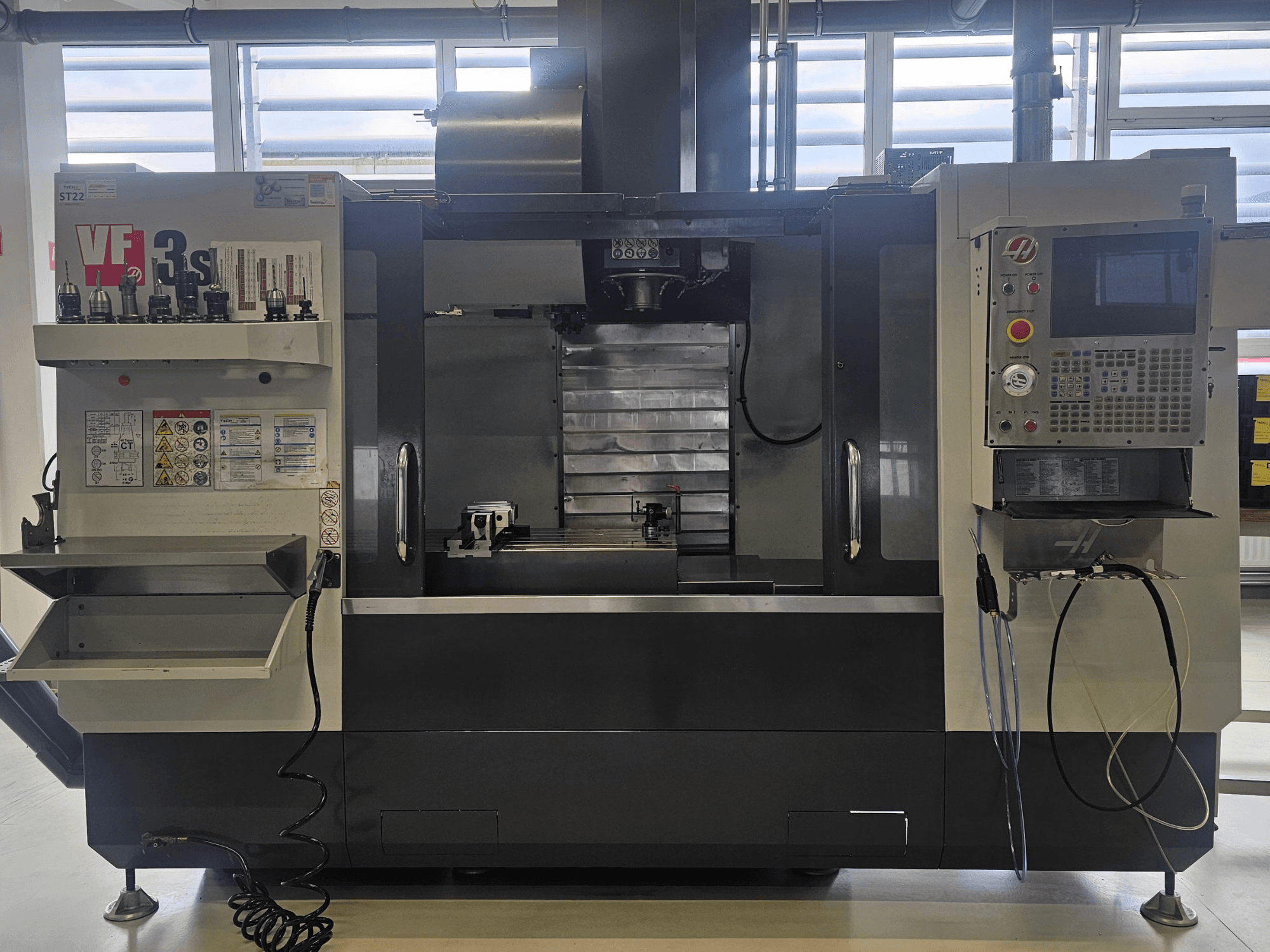 Haas VF3SS CNC mill, front view, featuring tool holders, control panel, and empty coolant tray, with safety labels visible.