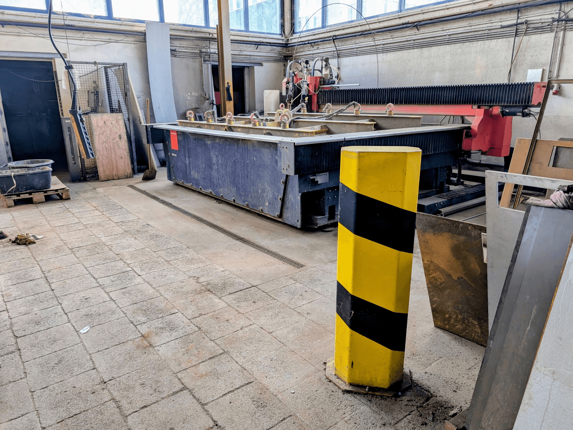 Bystronic Byjet L 2030 waterjet cutting machine in a workshop, side view, with a yellow safety pole and surrounding materials.