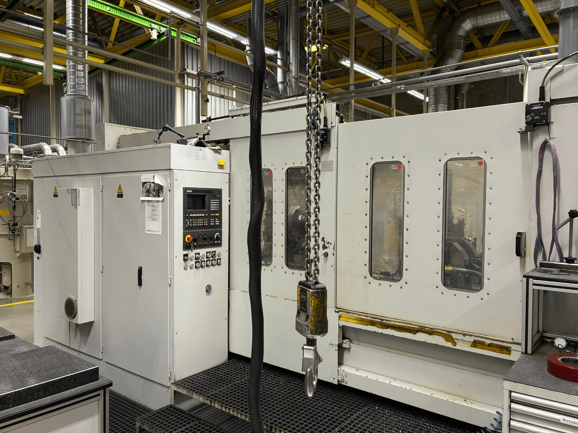 Industrial CNC machine with control panel, side view; features windows, chains, and metal components in a factory setting.