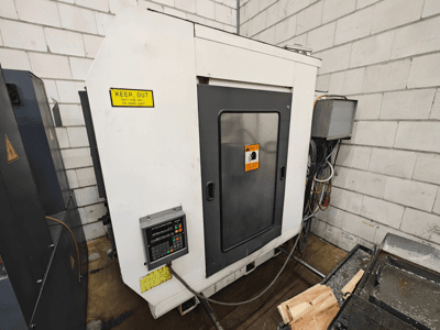 Industrial CNC machine, side view; features control panel, warning signs, and wood pieces on a work surface.